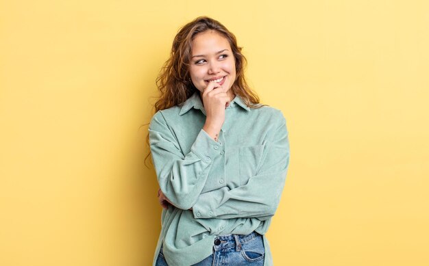 Hispanic pretty woman smiling with a happy, confident expression with hand on chin, wondering and looking to the side