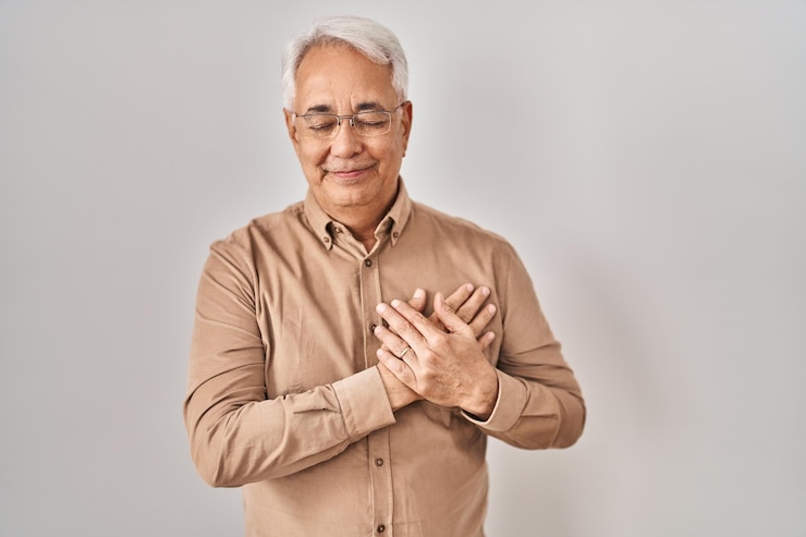 Photo 眼鏡をかけたヒスパニック系の年配の男性が、目を閉じて胸に手を当てて微笑み、顔の健康コンセプトに感謝のジェスチャーをする