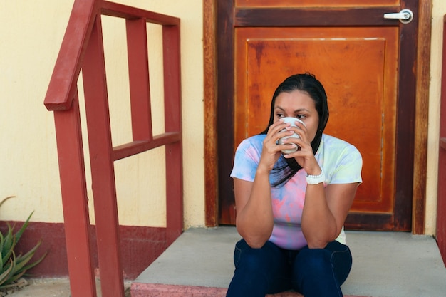 Photo hispanic woman drinking a cup of coffee sitting outside the house