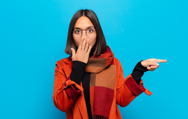 Hispanic woman feeling happy, shocked and surprised, covering mouth with hand and pointing to lateral copy space