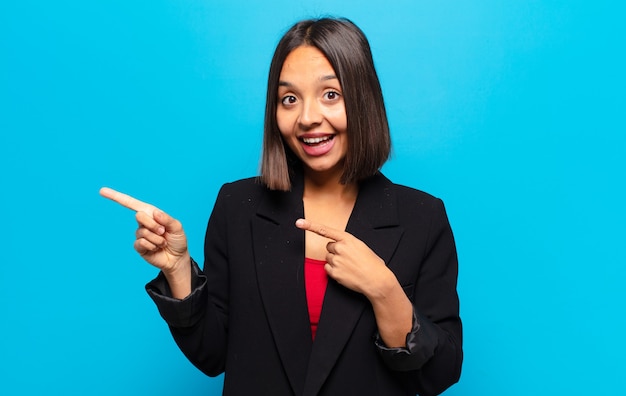 Hispanic woman feeling joyful and surprised, smiling with a shocked expression and pointing to the side