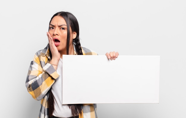 Hispanic woman feeling shocked and scared, looking terrified with open mouth and hands on cheeks