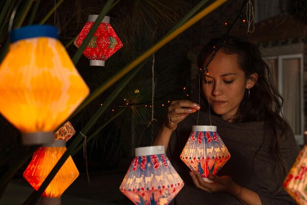 Hispanic woman holding a paper lantern while looking at it under a plant decorated with multiple lanterns