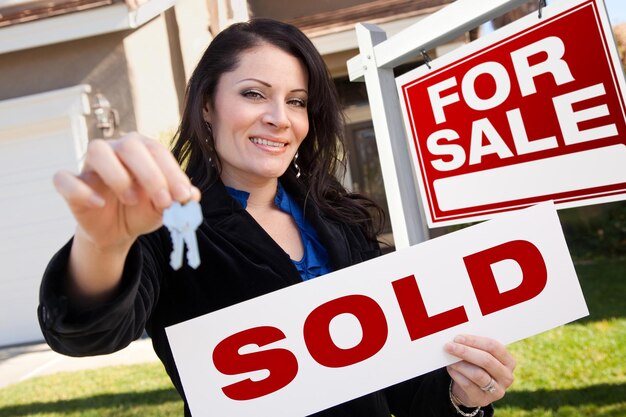 Photo hispanic woman holding sold real estate sign and keys in front house