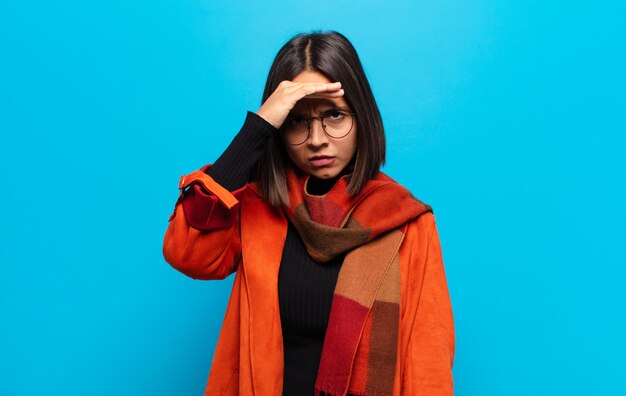 Hispanic woman looking bewildered and astonished, with hand over forehead looking far away, watching or searching