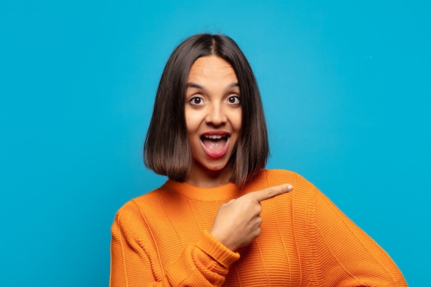 Hispanic woman looking excited and surprised pointing to the side and upwards to copy space