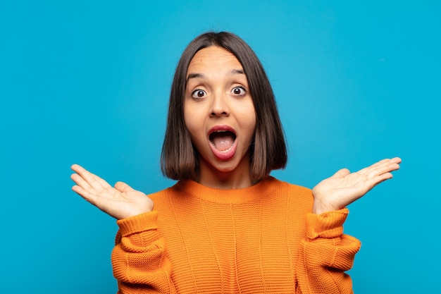 Hispanic woman looking happy and excited, shocked with an unexpected surprise with both hands open next to face