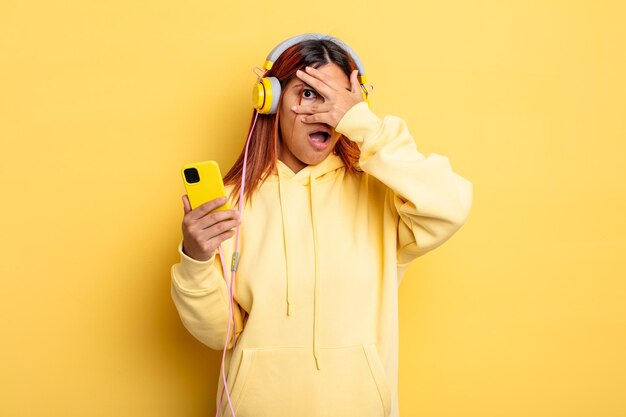 Hispanic woman looking shocked, scared or terrified, covering face with hand. headphones and smartphone concept