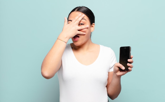 hispanic woman looking shocked, scared or terrified, covering face with hand and peeking between fingers