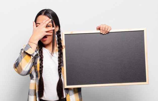 Hispanic woman looking shocked, scared or terrified, covering face with hand and peeking between fingers