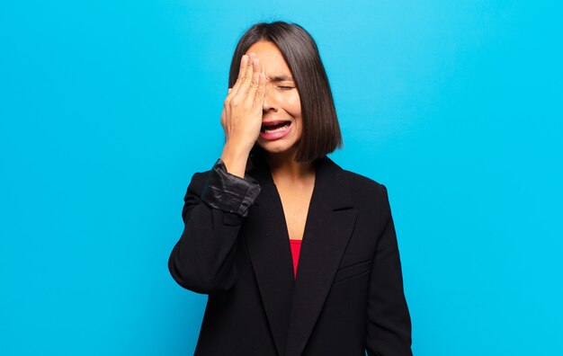 hispanic woman looking sleepy, bored and yawning, with a headache and one hand covering half the face