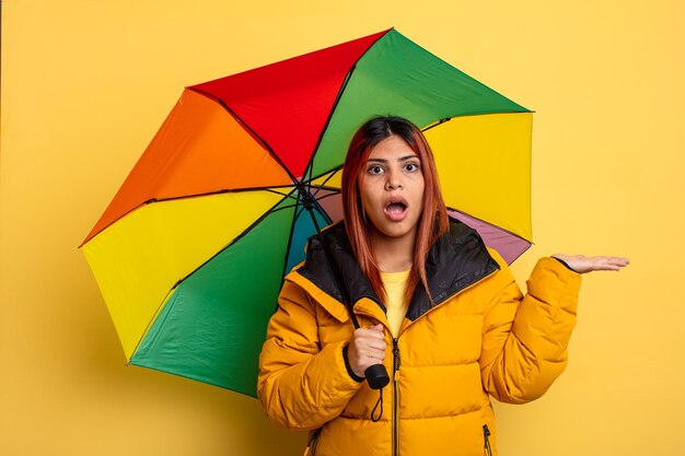 Hispanic woman looking surprised and shocked with jaw dropped holding an object rain and umbrella concept