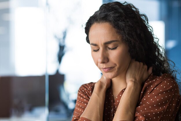 Una donna ispanica, stanca di lavorare in un ufficio moderno, una donna d'affari ha un forte dolore al collo che massaggia il collo