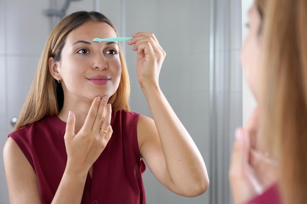 Hispanic woman shaving her eyebrows with razor in the mirror at home