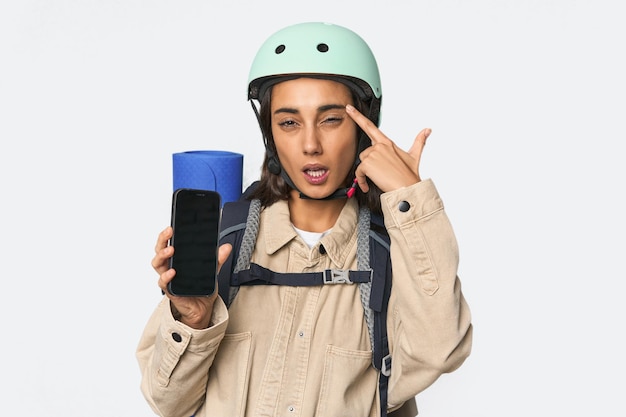 Hispanic woman shows phone with helmet showing a disappointment gesture with forefinger