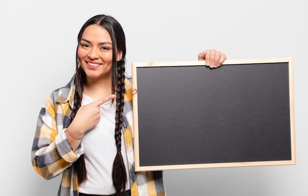 Hispanic woman smiling cheerfully, feeling happy and pointing to the side and upwards, showing object in copy space