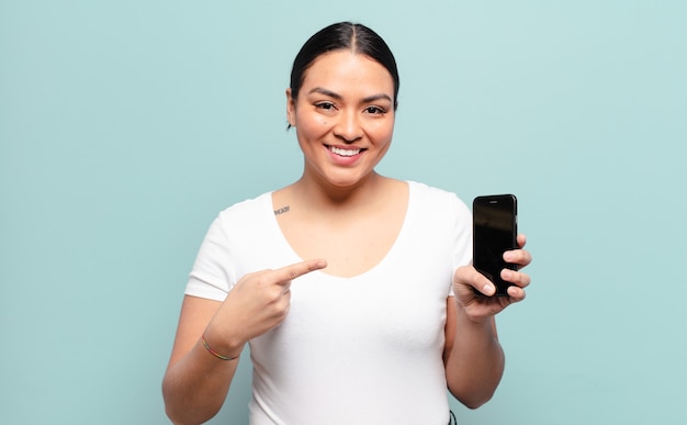 Hispanic woman smiling cheerfully, feeling happy and pointing to the side and upwards, showing object in copy space