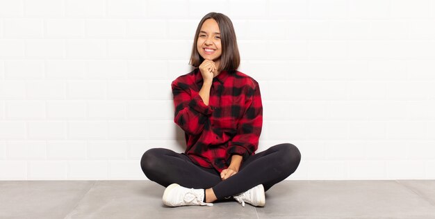 Hispanic woman smiling, enjoying life, feeling happy, friendly, satisfied and carefree with hand on chin