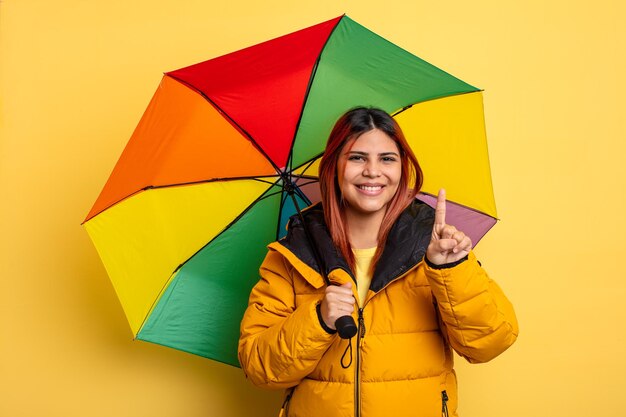 Hispanic woman smiling and looking friendly showing number one rain and umbrella concept