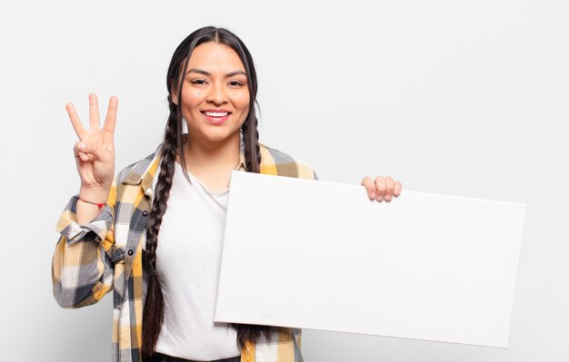 Hispanic woman smiling and looking friendly, showing number three or third with hand forward, counting down