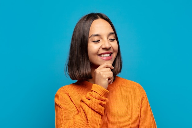 Hispanic woman smiling with a happy, confident expression with hand on chin, wondering and looking to the side