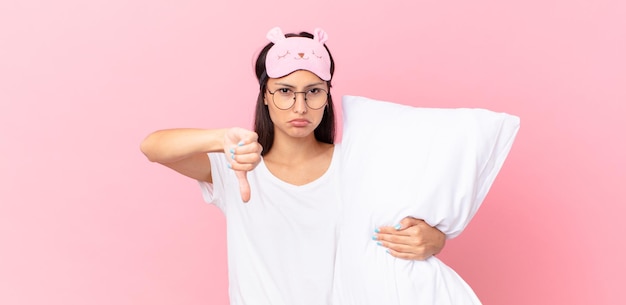 Hispanic woman wearing pajamas feeling cross,showing thumbs down and holding a pillow