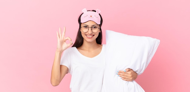 Hispanic woman wearing pajamas feeling happy, showing approval with okay gesture and holding a pillow