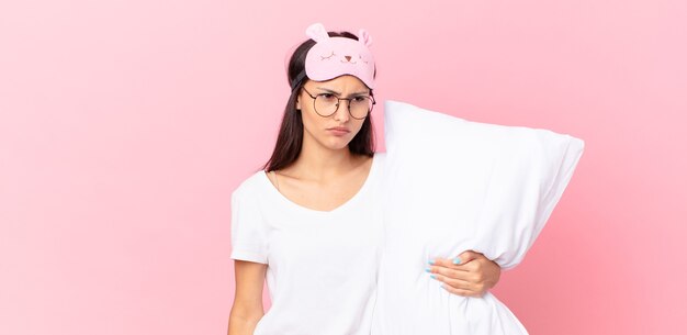 Hispanic woman wearing pajamas feeling sad, upset or angry and looking to the side and holding a pillow