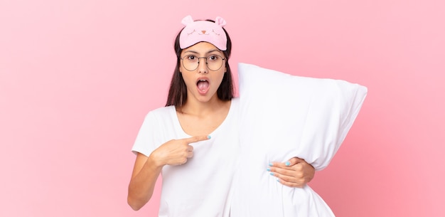 Hispanic woman wearing pajamas looking shocked and surprised with mouth wide open, pointing to self and holding a pillow