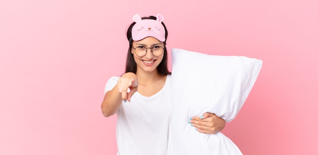 Hispanic woman wearing pajamas pointing at camera choosing you and holding a pillow