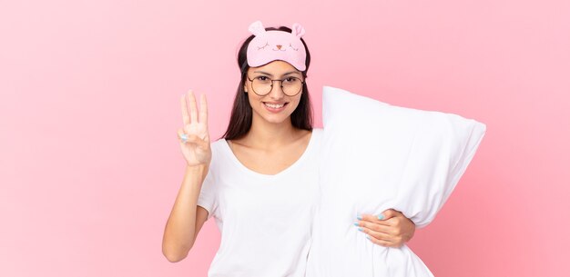 hispanic woman wearing pajamas smiling and looking friendly, showing number three and holding a pillow