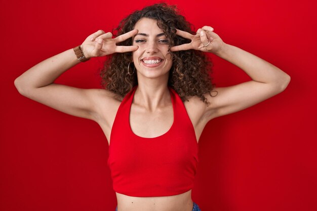 Hispanic woman with curly hair standing over red background doing peace symbol with fingers over face, smiling cheerful showing victory