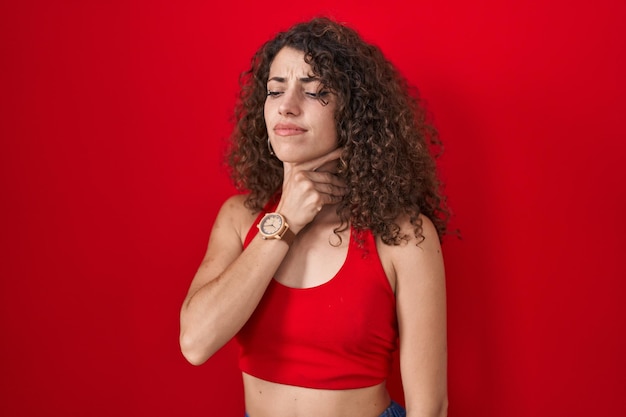 Hispanic woman with curly hair standing over red background touching painful neck sore throat for flu clod and infection