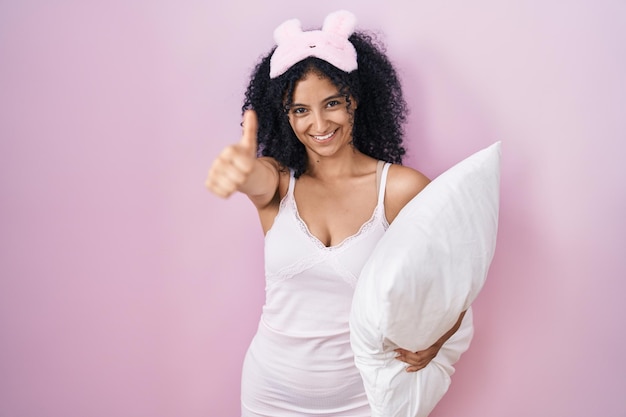 Hispanic woman with curly hair wearing sleep mask and pajama holding pillow approving doing positive gesture with hand thumbs up smiling and happy for success winner gesture