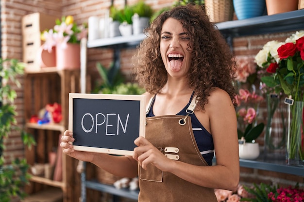 Foto donna ispanica con i capelli ricci che lavora al fiorista con cartello aperto che attacca la lingua felice con un'espressione divertente