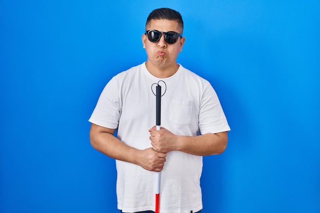 Hispanic young blind man holding cane puffing cheeks with funny face mouth inflated with air catching air