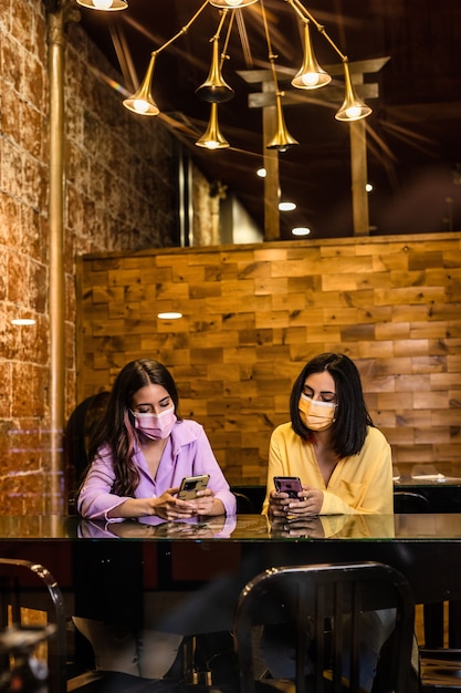 Giovani amiche ispaniche che utilizzano la tecnologia dello smartphone al ristorante per ordinare la cena e chiacchierare indossando la maschera per il viso stile di vita da donne indipendenti insieme colore giallo e lavanda 2021