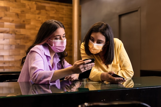 Hispanic young female friends using smartphone technology in restaurant to order dinner and chatting Wearing face mask Lifestyle from independent women together Yellow and lavender color 2021