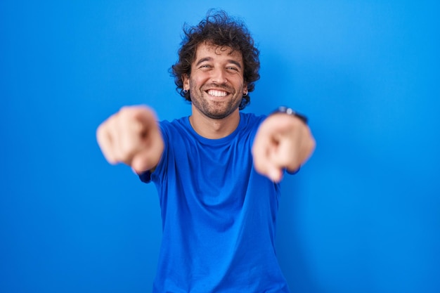 Испаноязычный молодой человек, стоящий на синем фоне, указывая пальцами на вас и камеру, улыбаясь позитивно и весело