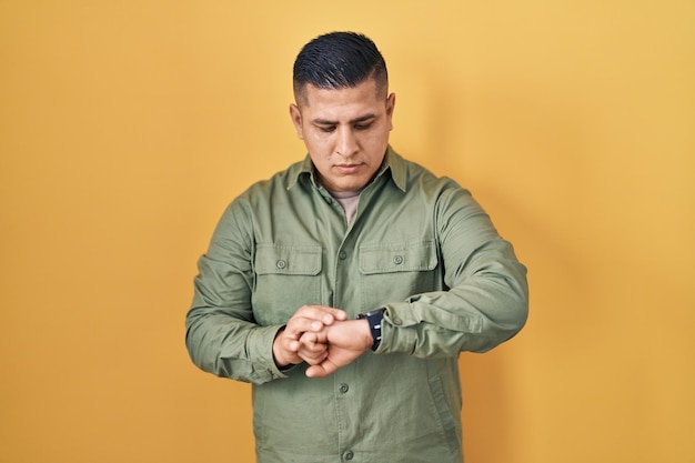 Hispanic young man standing over yellow background checking the time on wrist watch relaxed and confident