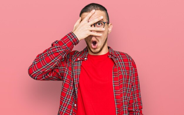 Hispanic young man wearing casual clothes peeking in shock covering face and eyes with hand looking through fingers with embarrassed expression