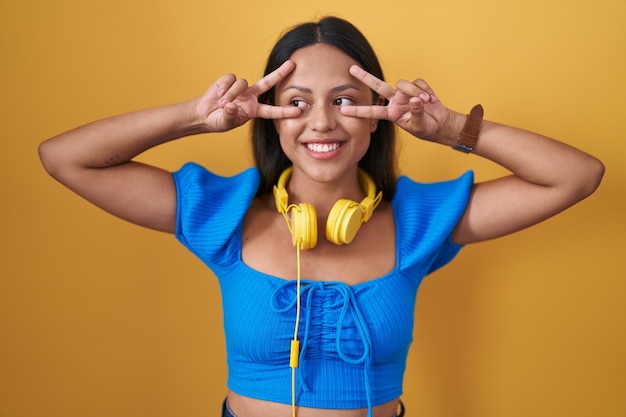 Hispanic young woman standing over yellow background doing peace symbol with fingers over face, smiling cheerful showing victory