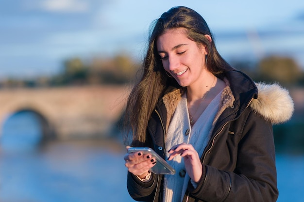 Hispanic young woman use smartphone for social networks near tajo river toledo winter cold style