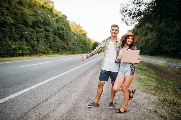 Hitchhiking couple holds blank cardboard with copy space. Hitchhike adventure of young man and woman. Happy hitchhikers on road