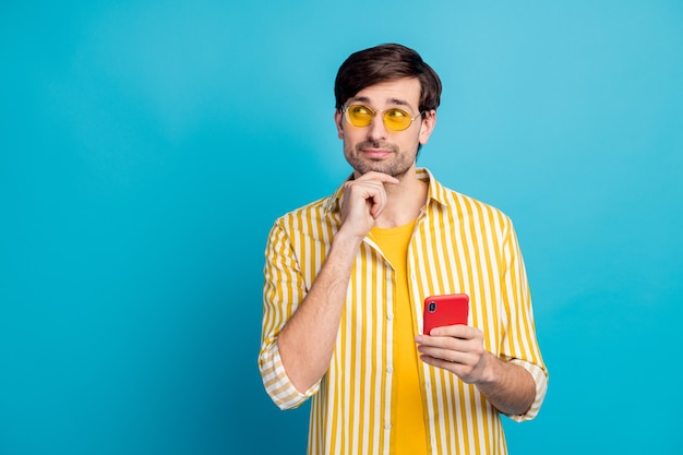 흠 여름 휴양지를 결정합니다. 마음이 있는 남자 여행자는 스마트폰 모양 카피스페이스를 사용하여 생각이 소셜 미디어 여행 광고를 선택한다고 생각합니다. 파란색 배경 위에 격리된 노란색 흰색 옷을 입습니다.