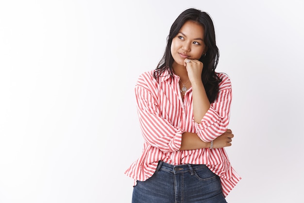 Hmm ziet er interessant uit. Portret van geïntrigeerde en nieuwsgierige aantrekkelijke jonge aziatische vrouw met tatoeage in gestreepte blouse dagdromen, leunend op de arm, wegkijkend naar de linkerbovenhoek met een dromerige glimlach