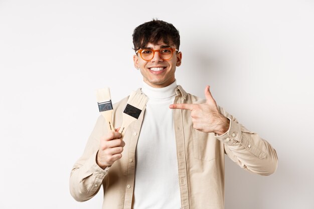 Hobbies and leisure concept. Cheerful young man in glasses pointing at painting brushes, renovating apartment, standing on white wall.