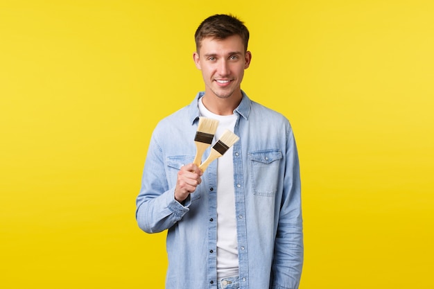 Hobbies, leisure and people lifestyle concept. Handsome smiling man suggesting renovate house, showing two painting brushes and grinning, learning how to draw, standing yellow background.