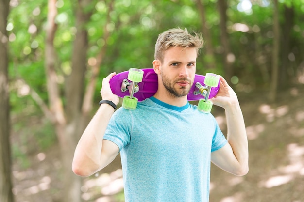 Hobby concept. Man smiling face posing with penny board park nature background, defocused. Guy carries penny board on shoulders, ready to ride. Man likes to ride skateboard and sporty lifestyle.