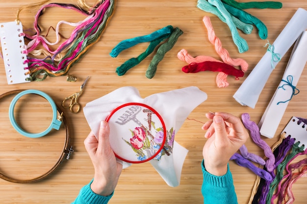 Hobby, artigianato, creatività, concetto di tempo libero a casa. mani femminili che ricamano a punto croce sulla tela. disposizione piatta, vista dall'alto